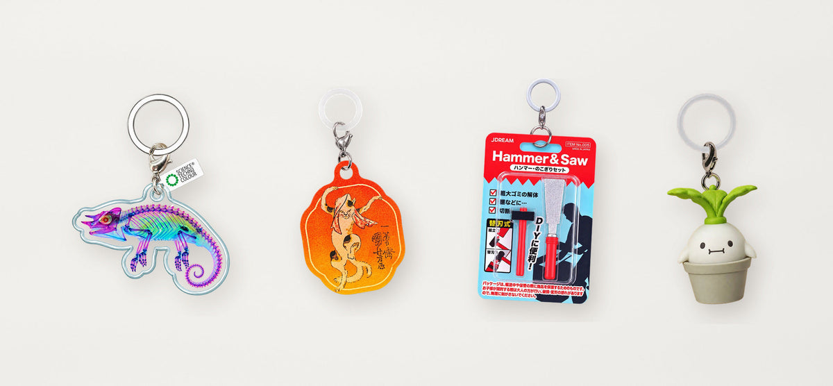 Four genuine Japanese miniature collectibles arranged on a neutral tabletop: transparent chameleon charm, ukiyo-e art charm, miniature toolkit, and mandrake character